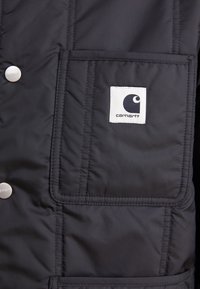 Schwarze Steppjacke mit ge quilteter Textur, ausgestattet mit einer großen Fronttasche und einem weißen Logo-Patch auf der Brust. Mit Druckknöpfen.