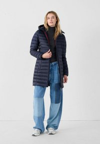 Marineblauwe pufferjas met een capuchon, gequilt textuur, voorrits en rode accenten, gecombineerd met wijde patchwork spijkerbroeken.