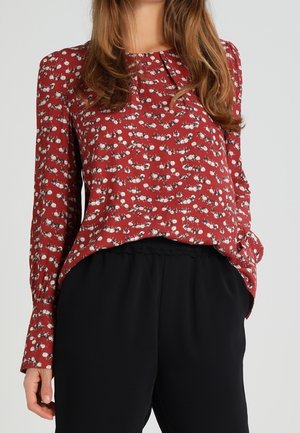 Femme portant un chemisier rouge à manches longues avec un motif floral blanc et un pantalon noir taille haute sur un fond uni.