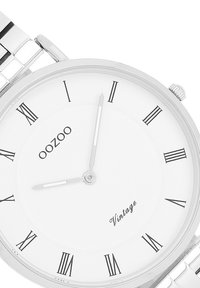 Montre analogique argentée avec un cadran blanc, des chiffres romains noirs et un bracelet métallique. Ses caractéristiques incluent un design simple et une petite couronne.