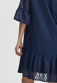 Laivansinis mekko, jossa on teksturoitu aalto-kuvio, lyhyet röyhelöhihet ja levenevä helma, jossa on päällysrakenne. Valmistettu kevyestä kankaasta.