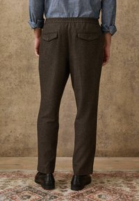 Brown wool blend pants with a relaxed fit, elastic waistband, and two back pockets with button closures, paired with black shoes.