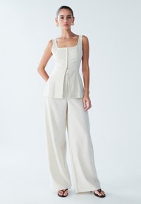 Licht beige mouwloos topje met vierkante halslijn en plooien aan de voorkant, gecombineerd met wijde broek gemaakt van gestructureerde stof. Zwarte flip-flops.