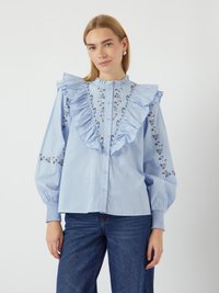 Blusa azzurra chiara con colletto a balze, ricami floreali sulle maniche e chiusura frontale a bottoni. Il tessuto è morbido con maniche a palloncino.