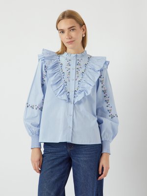 Light blue blouse with a ruffle collar, floral embroidery on sleeves, and button front closure. Fabric is smooth with puffed sleeves.