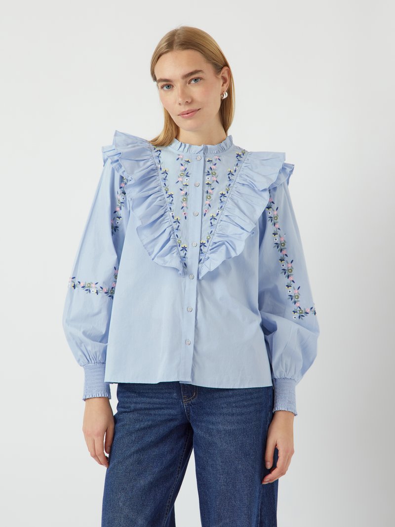 Blusa azzurra chiara con colletto a balze, ricami floreali sulle maniche e chiusura frontale a bottoni. Il tessuto è morbido con maniche a palloncino.