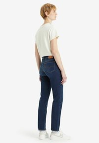 Jeans de mezclilla azul oscuro con cintura alta y corte ajustado, combinados con una camiseta de manga corta color crema y zapatillas blancas. Parche de marca visible en la cintura.