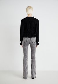 Blouse noire à volants avec des manches longues, assortie à un jean gris évasé présentant une texture déchirée et des coutures contrastantes visibles sur les poches.