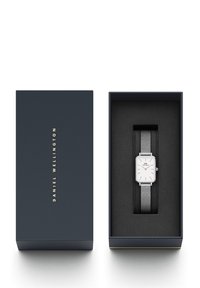 Montre rectangulaire en argent avec bracelet en maille, présentée dans une boîte bleu marine. L'intérieur de la boîte est noir, avec un logo sur le cadran de la montre et sur la boîte.