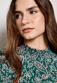 Tee-farbene Bluse mit floralem Muster, einem gerafften hohen Kragen und zarten weißen und beigen Blumenmotiven. Weicher, leichter Stoff mit einer strukturierten Oberfläche.