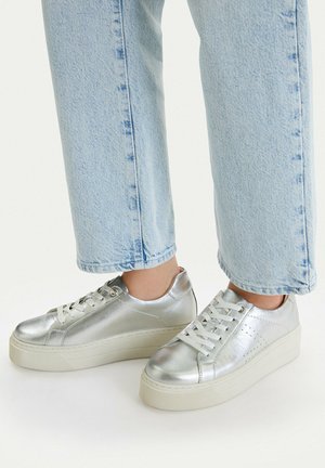 Nohy v stříbrných metalických teniskách na platformě spárované se světle modrými zkrácenými džínami na bílém pozadí.