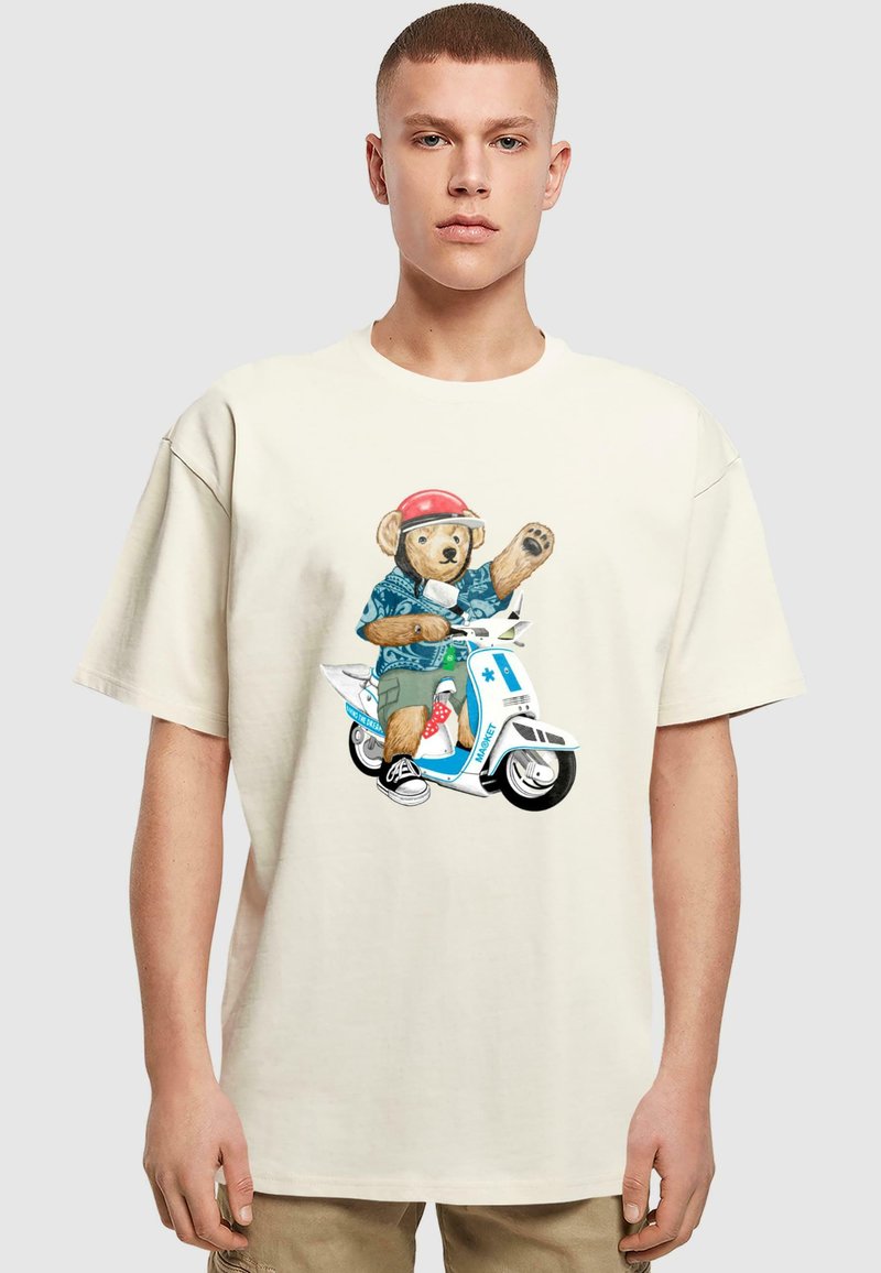 Camiseta de gran tamaño color crema que presenta un gráfico de un oso en un patinete, con un casco rojo, camisa azul, pantalones cortos verdes y zapatos blancos.
