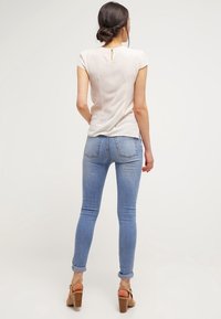 Světle béžový top s krátkým rukávem a klíčovou dírkou na zádech, kombinovaný se světle modrými skinny džínami, které mají hladkou strukturu a přiléhavý design.