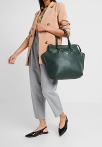 Kobieta ubrana w beżową marynarkę, kwiecistą koszulę, szare spodnie oraz czarne buty na obcasie typu slingback, niesie na ramieniu dużą ciemnozieloną torbę.