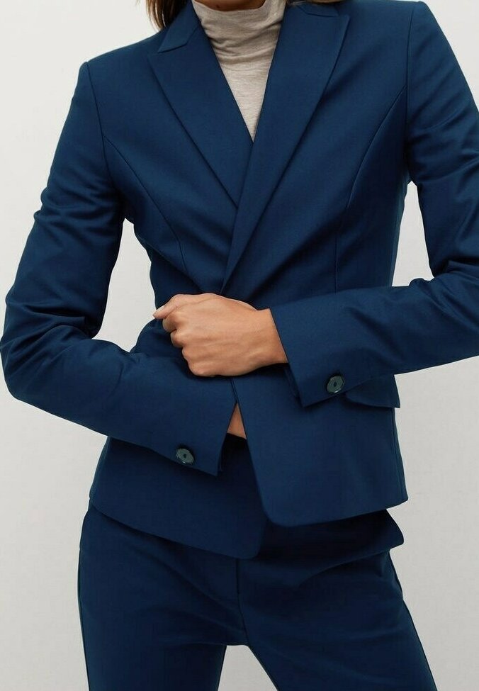 Veste bleu marine sur mesure avec un col cranté, un tissu texturé, des poches avant et des poignets boutonnés, associée à un col roulé gris.