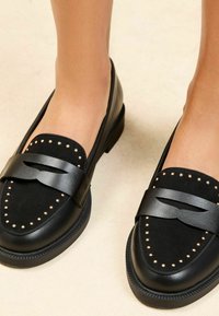 Loafers negri din piele şi ante, cu un design cu studuri, curea lată şi talpă din cauciuc, prezentate dintr-o perspectivă unghiulară.