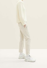 Personne portant un pull côtelé couleur crème, un pantalon beige slim, des chaussettes blanches et des baskets blanches épaisses, debout de profil avec les mains dans les poches.