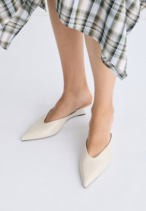 Femme portant des mules crème à bout pointu et une jupe à carreaux, debout sur un sol blanc.