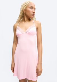 Junge Frau mit langen blonden Zöpfen, die ein hellrosa Nachthemd mit Spitzenbesatz trägt und nach rechts blickt, vor schlichtem Hintergrund.