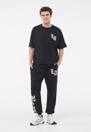 T-shirt in cotone nero con grafica bianca "LA" sul lato sinistro del petto, abbinato a pantaloni felpati neri con testo "LA" e "BASIC", indossati con sneakers bianche.