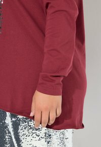 Bordeaux langekleurig shirt met een gebogen zoom en een zachte textuur, gecombineerd met een glanzende zilveren paillettenrok. Hand steunt op de rand van het shirt.