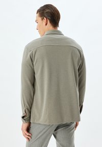 Olivgröna långärmade skjorta med en texturerad yta, knäppt manschetter och avslappnad passform. Förstärkt sömdetalj längs axlarna.