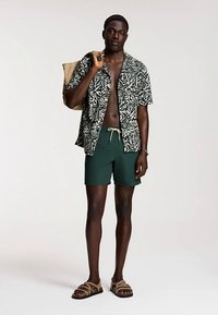 Koszula z krótkim rękawem w czarno-białe wzory, zielone szorty, beżowe sandały, trzyma beżową torbę na ramię. Charakteryzuje się casualowym stylem plażowym.