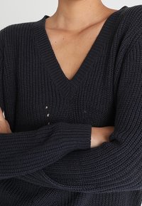 Pull en tricot bleu marine foncé avec un décolleté en V, texture côtelée et manches longues. Présente de petits détails de déchirure sur le devant.