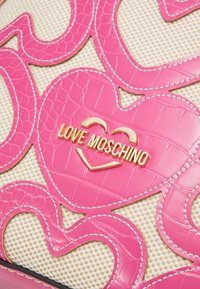 Rosa läderhandväska med hjärtdesign, guld "Love Moschino"-logotyp och en texturerad beige bakgrund med vita sömnadsdetaljer.