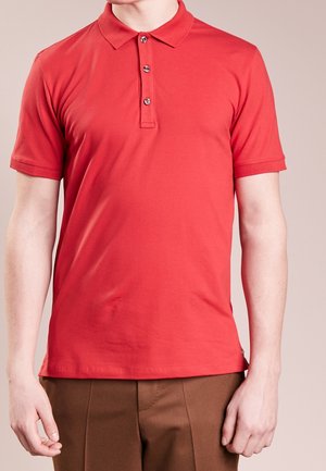 Mann trägt rotes Polohemd mit kurzen Ärmeln und drei Knöpfen sowie braune Hose, steht vor beigem Hintergrund.