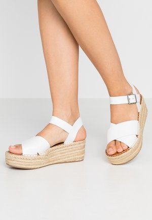 Sandales compensées blanches avec une semelle en espadrille, dotées de larges brides croisées et d'une fine bride de cheville avec une boucle argentée.