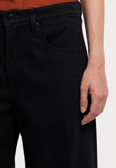 Jeans de ganga preta com corte reto, apresentando um bolso frontal, costura em contraste e fecho com botão de metal. Textura suave.