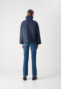 Ναυτικό καπιτονέ μπουφάν με ψηλό γιακά, απεικονιζόμενο από πίσω, σε συνδυασμό με τζιν ίσιας γραμμής σε μπλε χρώμα και μαύρες πλατφόρμες μποτών.