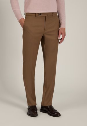Uomo che indossa pantaloni marroni su misura con scarpe eleganti nere in pelle e una camicia rosa chiaro a maniche lunghe, in piedi davanti a uno sfondo neutro.