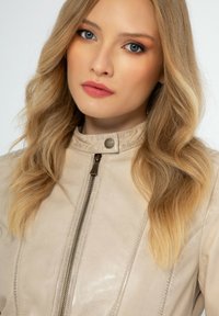 Béžová kožená bunda s vysokým límcem, předním zapínáním na zip a viditelnými švy. Hladká textura s minimalistickým designem.