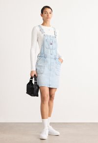 Lys blå denim overallkjole med frontlommer, justerbare stropper og en liten rød logopatch. Brukt over en hvit langermet skjorte.