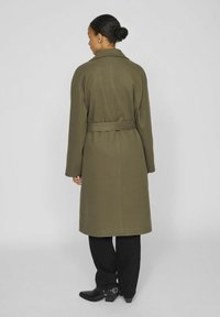 Olive zaļa mētelis ar jostu, apkakli, garām piedurknēm un taisnu griezumu. Valkāts virs melnām biksēm un potīšu zābakiem, ar gludu tekstūru.