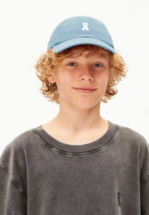 Junge mit lockigem roten Haar, der eine blaue Kappe und einen ausgewaschenen schwarzen Rundhalsausschnitt trägt, leicht lächelnd vor einem schlichten weißen Hintergrund.