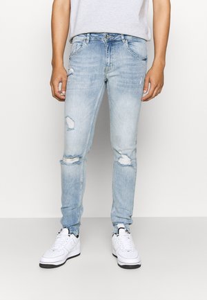 Mann trägt hellblaue, zerrissene Skinny-Jeans und weiße Sneakers, steht vor einem einfarbigen hellen Hintergrund.