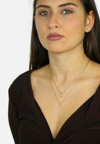 Collares en tonos dorados en capas que presentan un pendant rectangular y uno redondo, llevados sobre una blusa marrón de escote en V, con una textura suave y un acabado brillante.