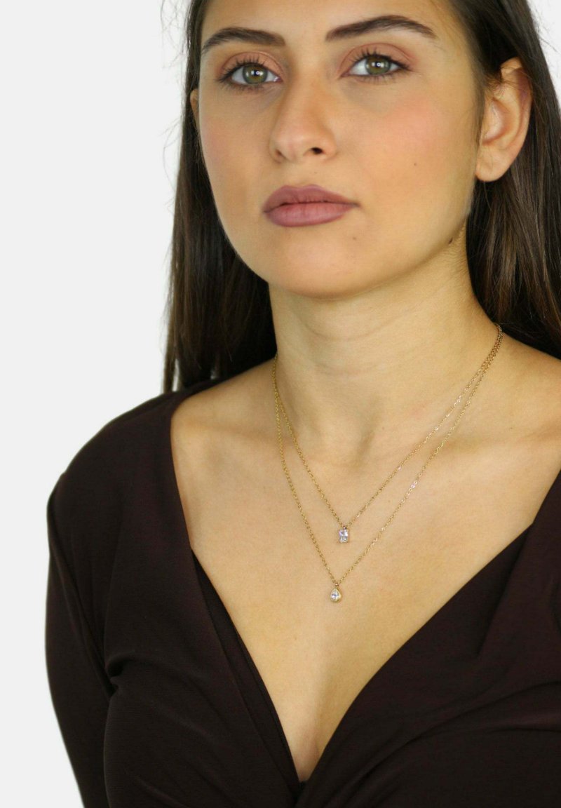 Collares en tonos dorados en capas que presentan un pendant rectangular y uno redondo, llevados sobre una blusa marrón de escote en V, con una textura suave y un acabado brillante.