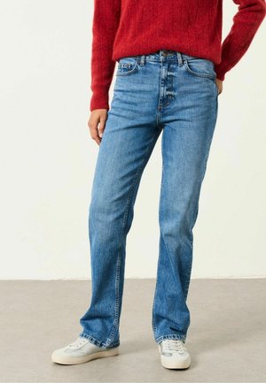 Persona que lleva vaqueros azul claro de corte recto, zapatillas blancas y un suéter rojo, de pie con una mano en el bolsillo.