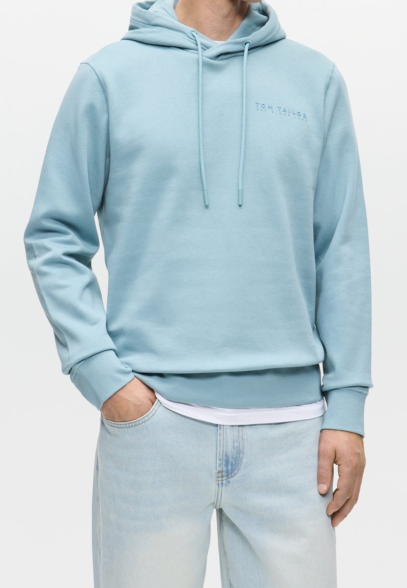 Homme portant un sweat à capuche bleu clair avec cordons et un jean bleu clair, main dans la poche, sur un fond uni.