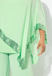Completo verde chiaro con una blusa trasparente e orlo di paillettes verdi scintillanti. Le maniche sono fluide, creando un effetto a strati. Pantaloni aderenti sotto.