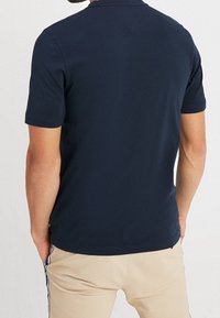 Mand i navy blå kortærmet polo shirt og beige bukser med sidestropper, stående med hænderne i lommerne og vendt den anden vej.