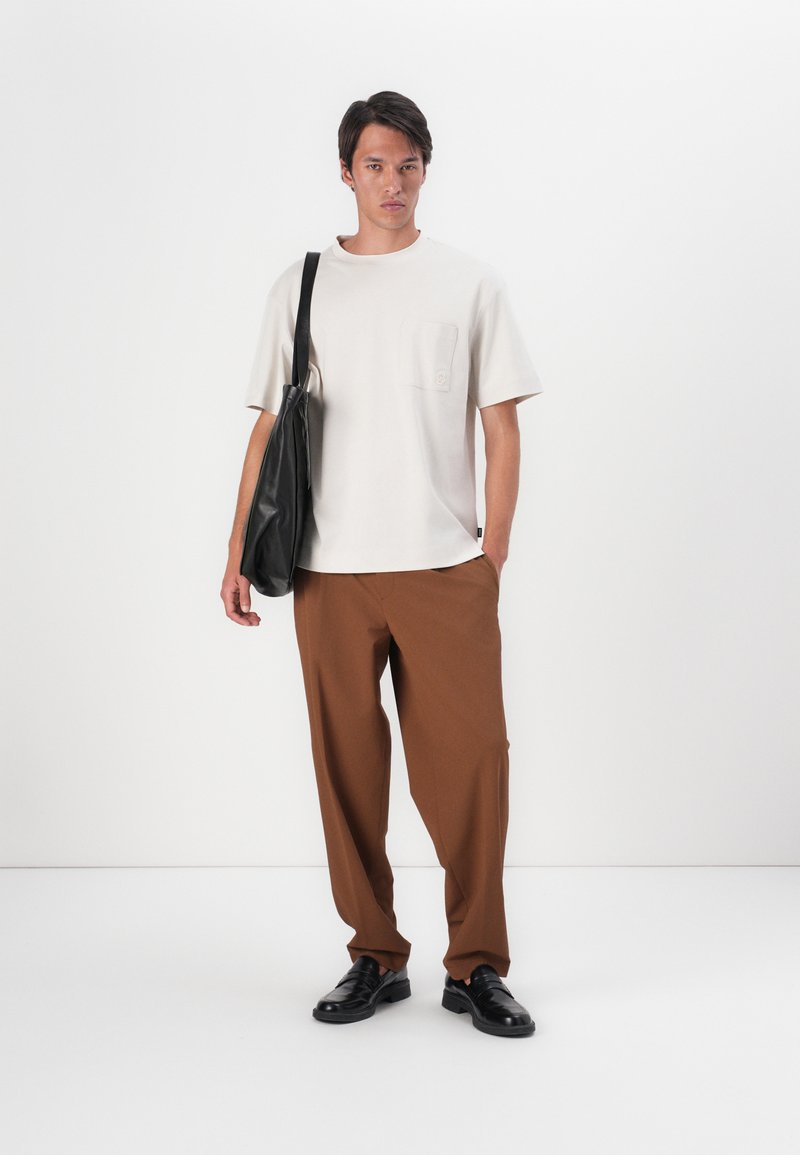 Jasnobeżowa koszulka z krótkim rękawem z kieszenią na piersi, w połączeniu z luźnymi brązowymi spodniami i czarnymi skórzanymi butami, trzymając czarną torbę na ramię.