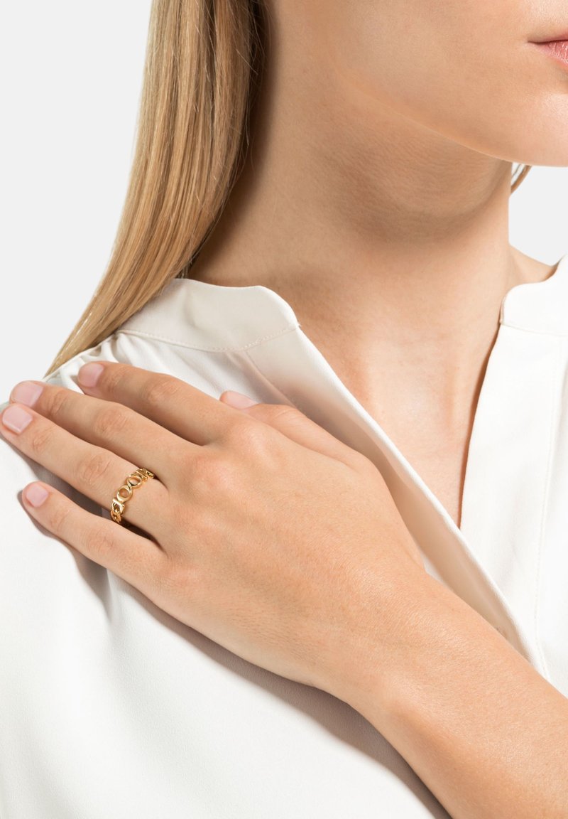 Anello d'oro con anelli circolari intrecciati e maglie. Indossato su una mano appoggiata su una camicetta bianca, che mette in risalto la pelle texturizzata e le unghie.