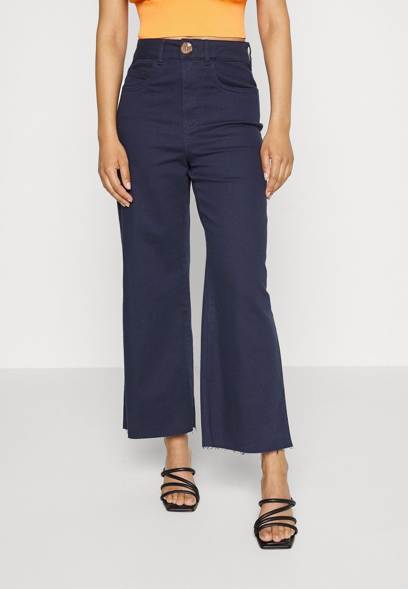 Vidje jeans i marinblå färg med rå kant, framfickor, knappstängning och åtsittande midja, i kombination med svarta klacksandaler med band.