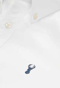 Weißes Hemd mit Knopfverschluss aus Baumwolle, versehen mit einem kleinen blauen gestickten Hirsch-Logo auf der Brusttasche. Weicher Stoff und klassischer Kragen.