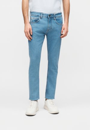 Muž nosi svetlomodré slim fit džínsy s prednými vreckami a biele tenisky, stojí pred jednofarebným svetlým pozadím.